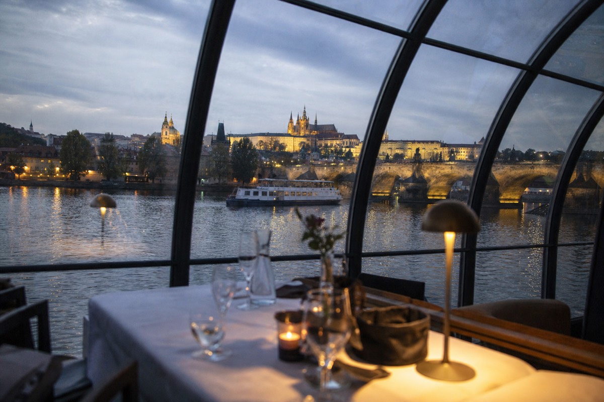 Dinner table set on a glass cruise boat with the lit Prague embankment behind