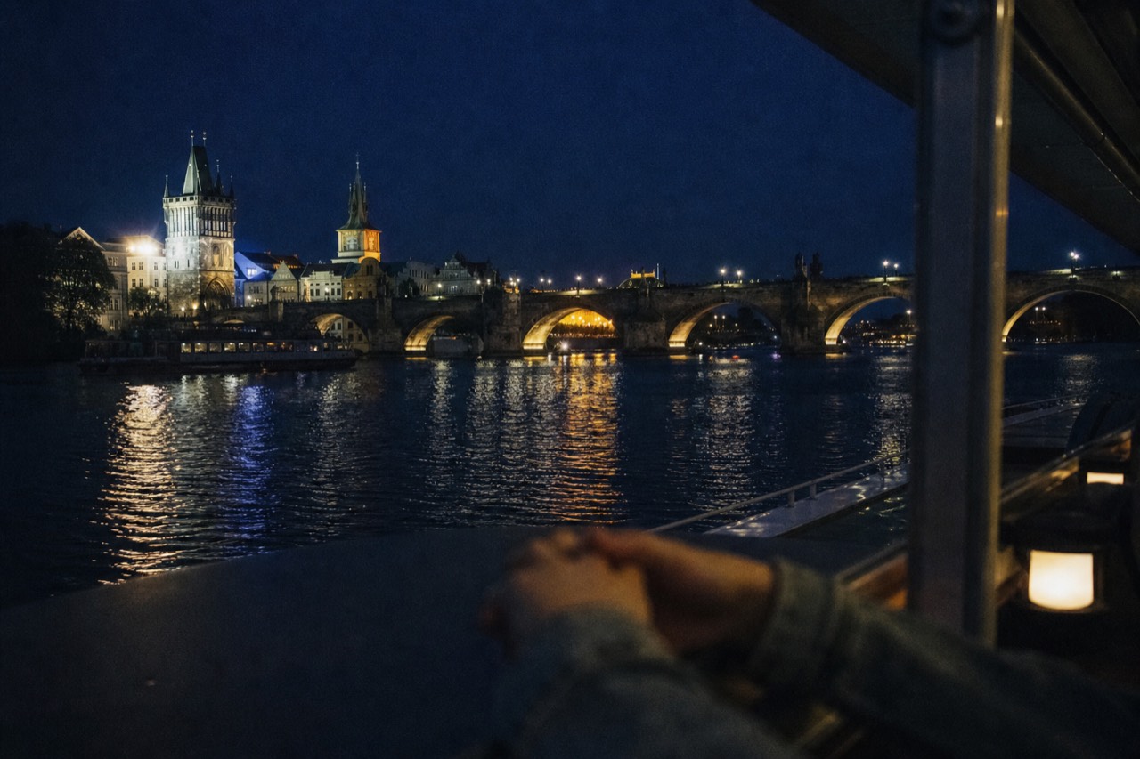 Open-top glass sightseeing boat passing under the floodlit Charles Bridge on the Vltava at night with Prague Castle glowing on the hill behind
