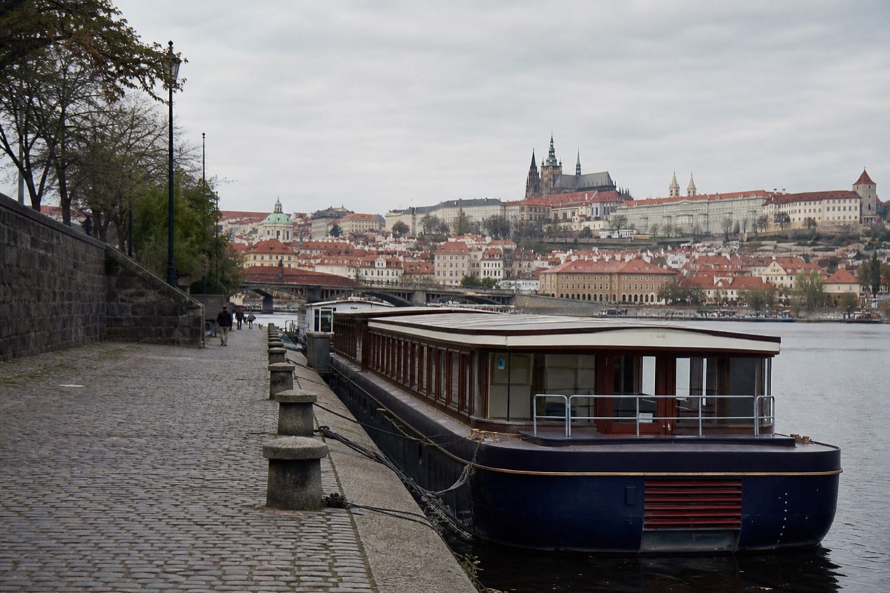 A red open-top sightseeing bus on a Prague street with the Vltava river and the castle hill visible in the background