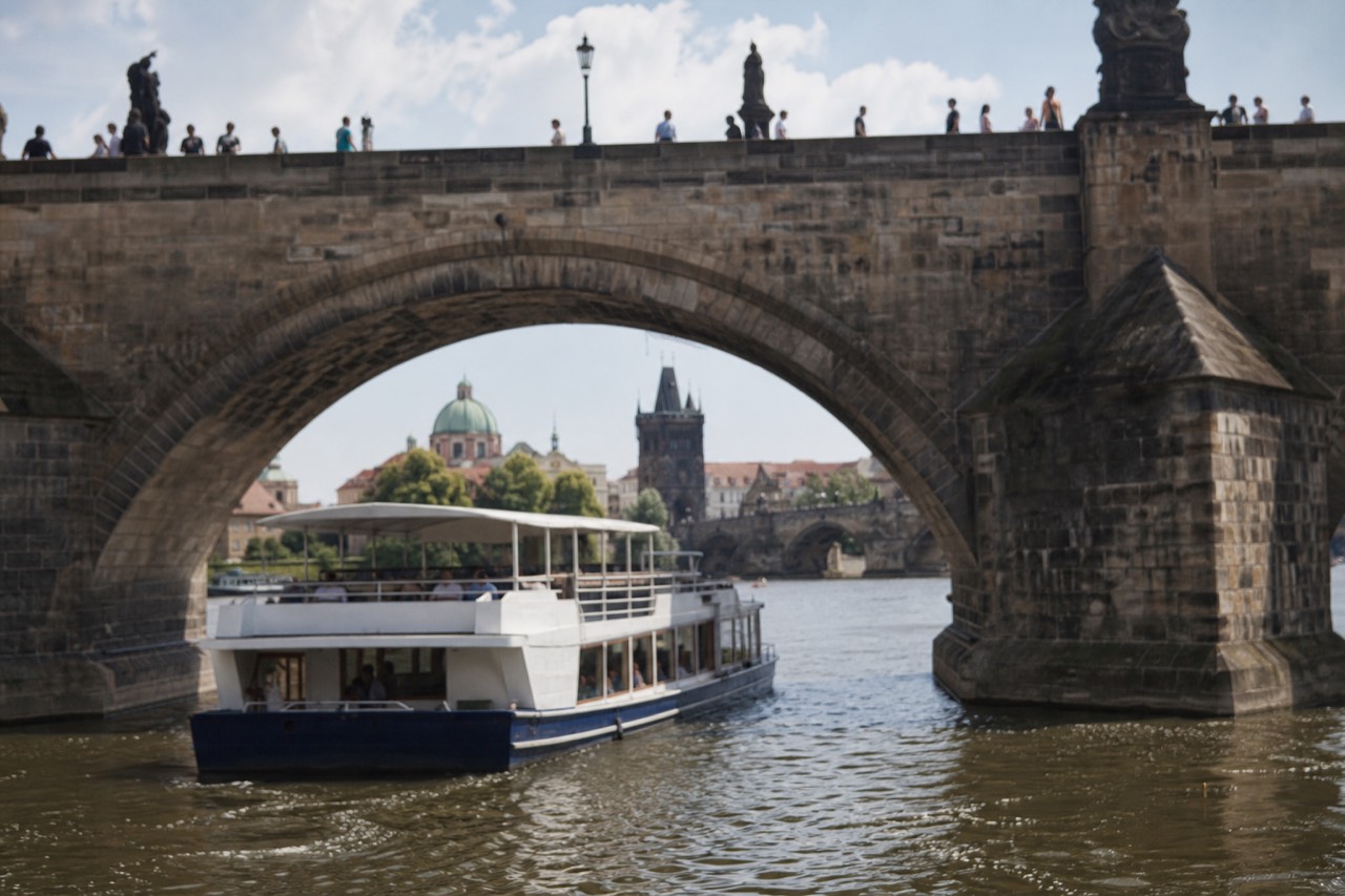 View over Prague's Old Town red rooftops toward the castle and the Vltava river with a sightseeing boat on the water below