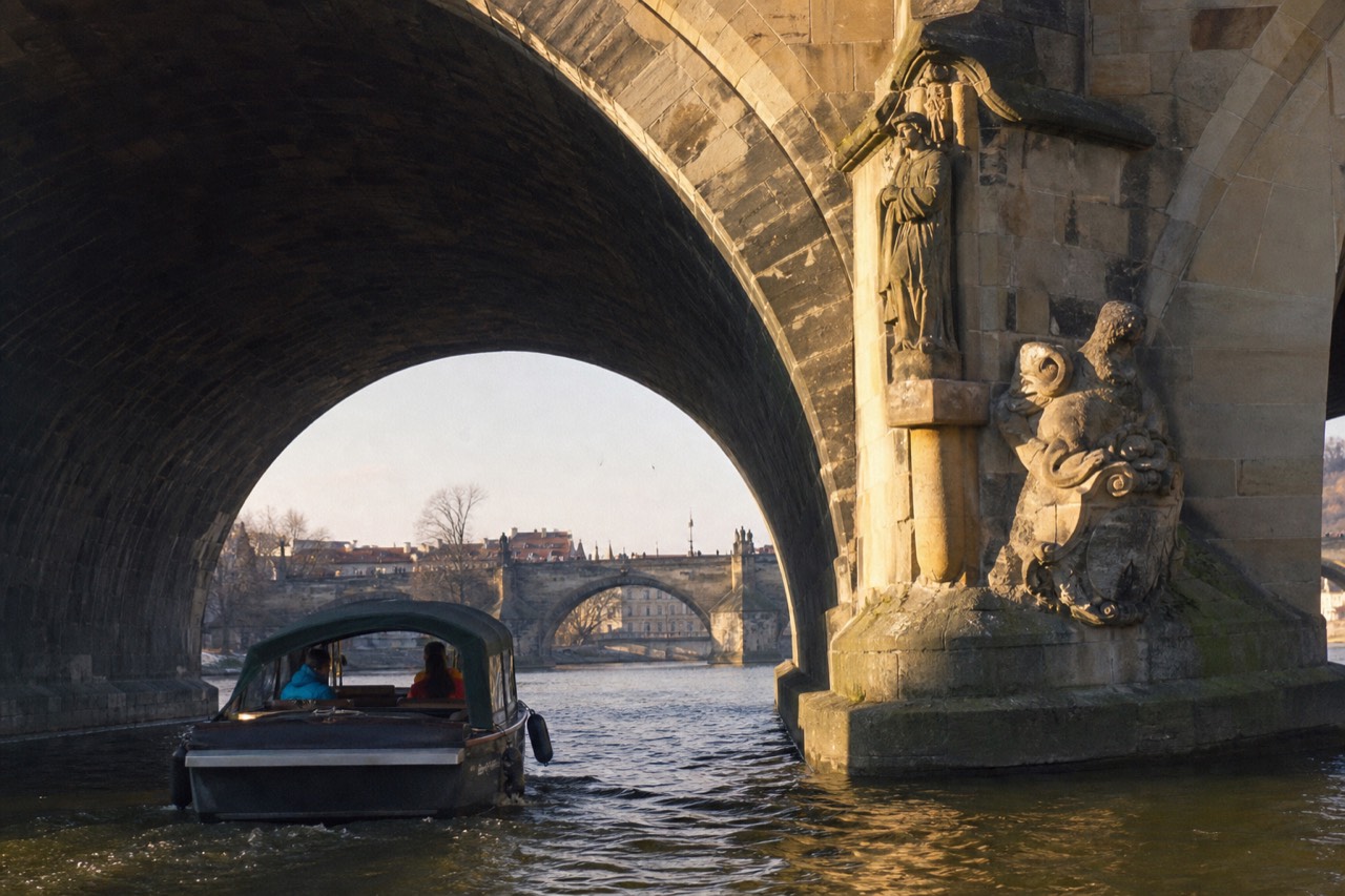 Charles Bridge and the floodlit Prague Castle seen from a boat on the Vltava at dusk, statues visible from below