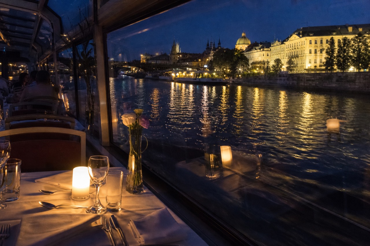 Set dinner tables inside an open-top glass boat on the Vltava at night, the floodlit Charles Bridge and Prague Castle passing behind the glass