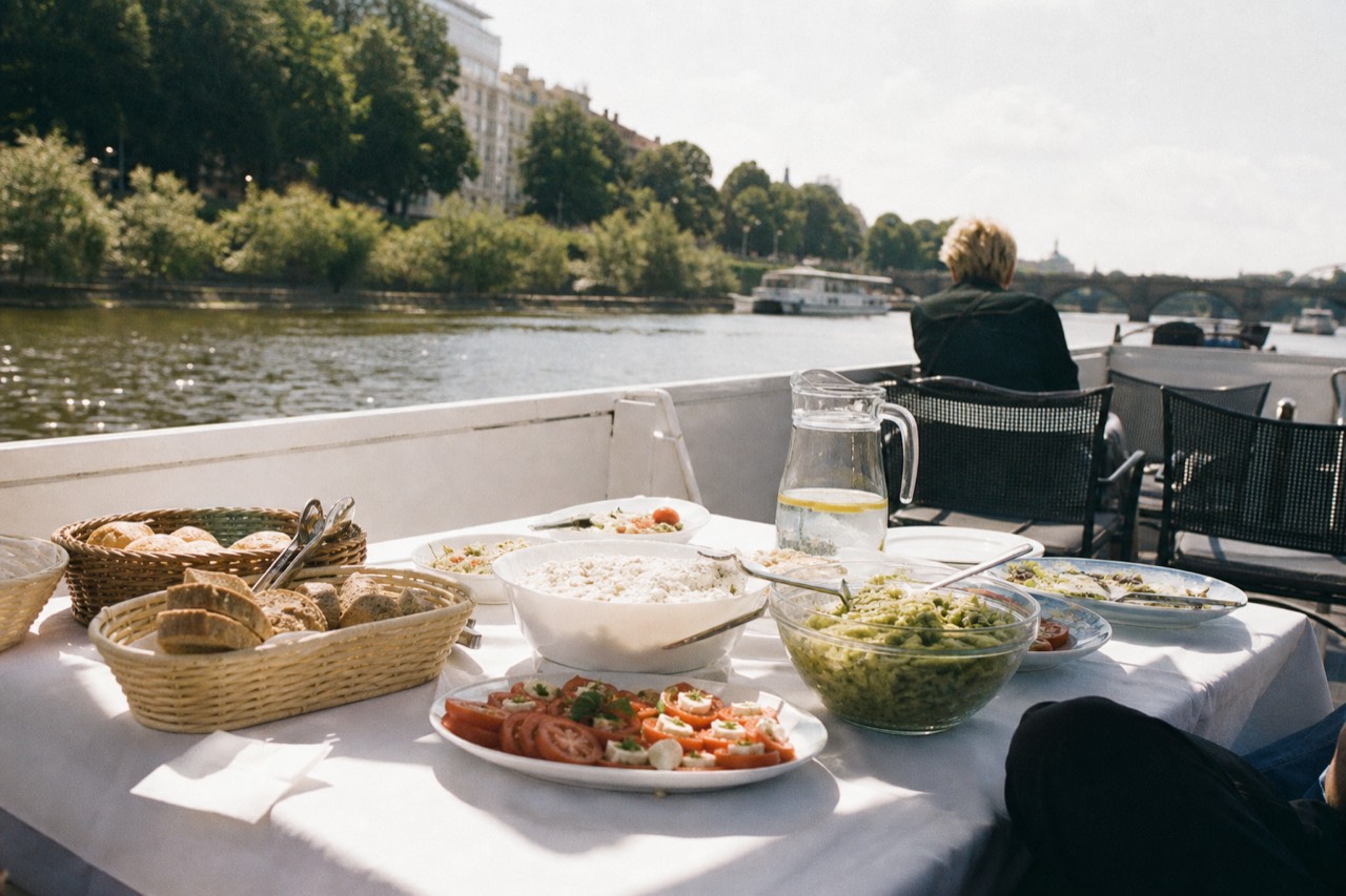 Open-top glass lunch boat on the Vltava in bright daylight, tables laid inside, Charles Bridge and Prague Castle in full colour behind it