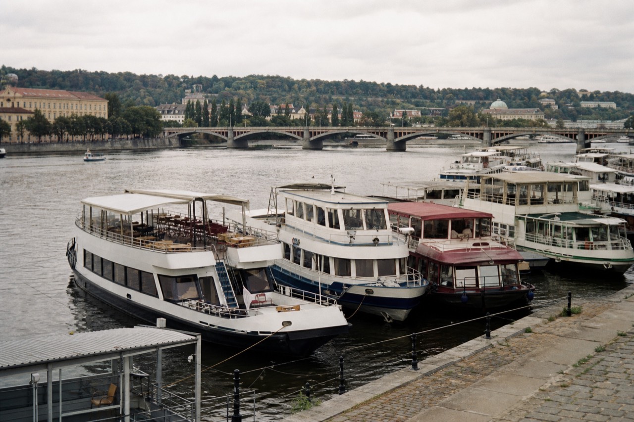 Passengers on the open deck of a Vltava sightseeing boat in daylight with the Prague skyline and river ahead