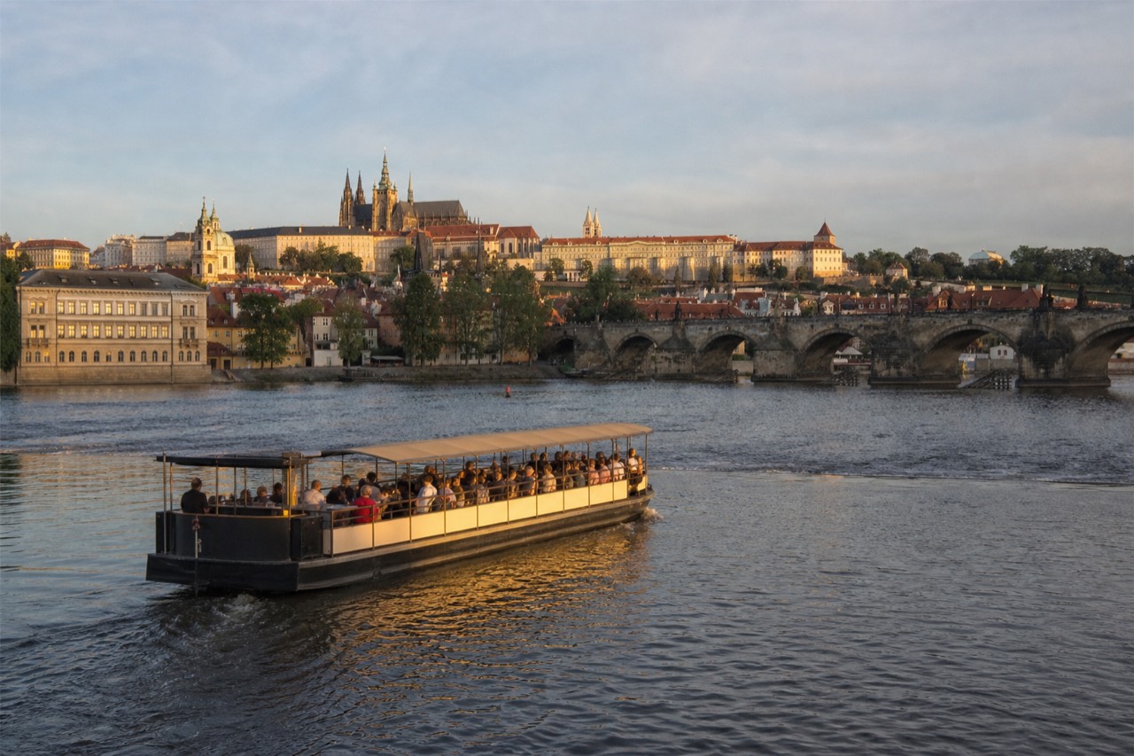 Open-top glass sightseeing boat on the Vltava passing under Charles Bridge with Prague Castle lit behind it at dusk