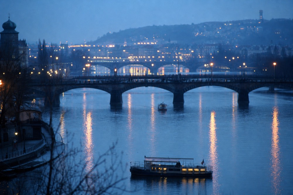 Several lit bridges receding into blue-hour haze along the Vltava, long reflections on calm water