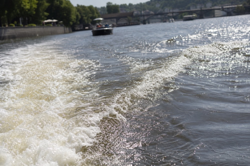 A boat's wake fanning out across the Vltava, sunlit foam and ripples filling most of the frame, the riverbank a soft blur at the edge