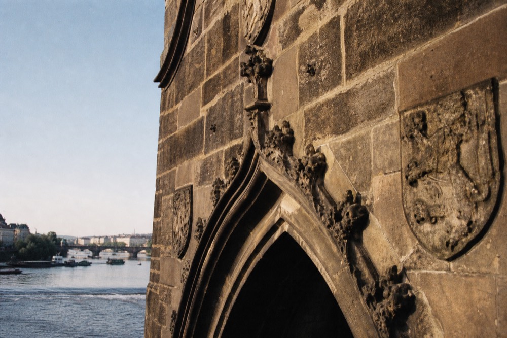The gothic stonework on one of Charles Bridge's towers, seen looking up from the water, weathered carved stone against a clear sky