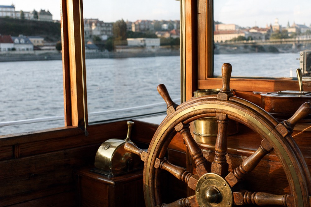 A boat's wooden helm wheel and brass fittings in soft window light, no markings or nameplates visible
