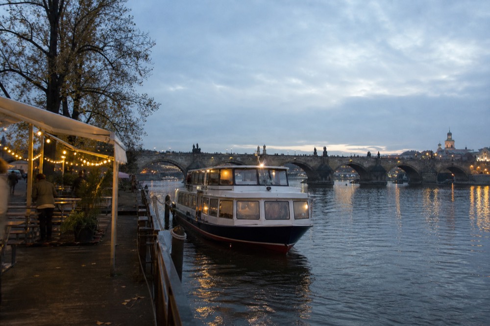 The Čechův most pier at dusk, strings of warm lights along the dock, a boat easing in to board