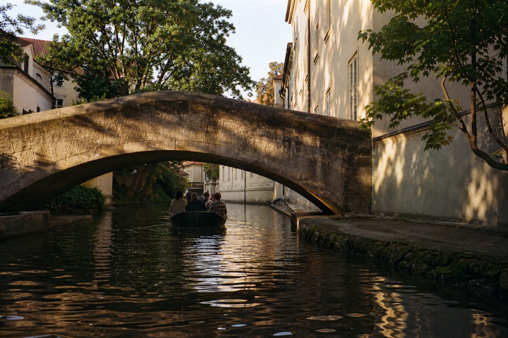 A small wooden boat gliding through the Čertovka canal, passing under a low stone footbridge