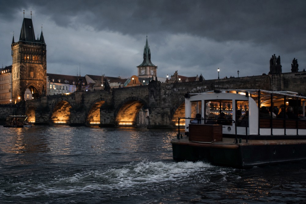 Charles Bridge at blue hour, warm lamplight glowing under the stone arches, a boat's wake crossing the foreground