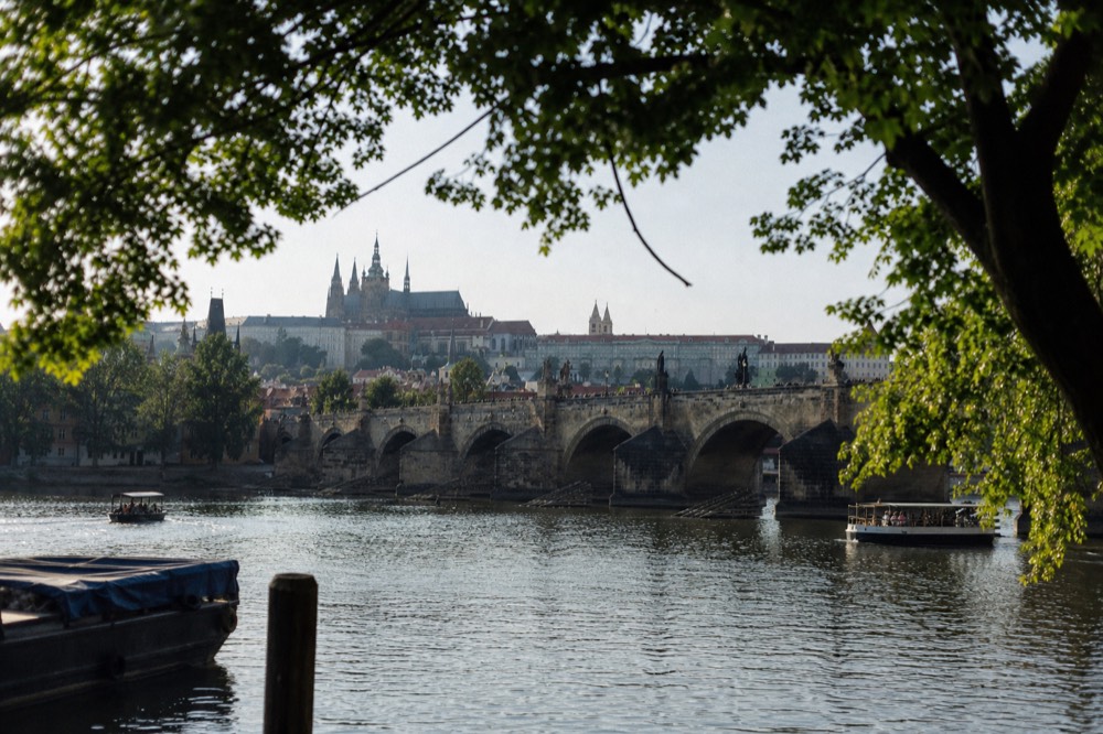 Charles Bridge seen low from the water in soft morning light, stone arches and the castle hill rising behind