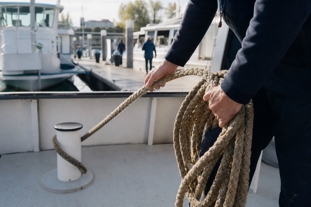 A crew member's hands coiling a mooring rope on a cruise boat's deck before departure, sunlit rope fibres in sharp focus, the pier soft behind