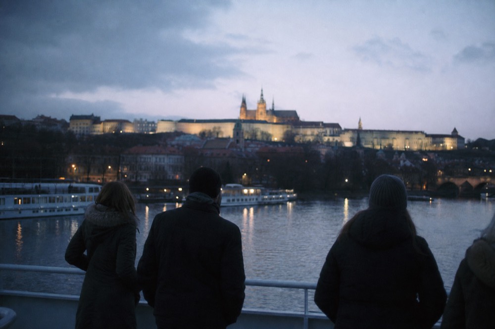 Passengers' backs at a boat's deck rail, looking toward the lit castle hill at dusk, distant and unidentifiable