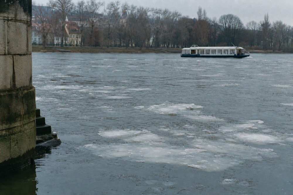 Small ice floes drifting slowly on the winter Vltava, the far bank bare of leaves under a pale overcast sky, a heated covered boat passing in the distance