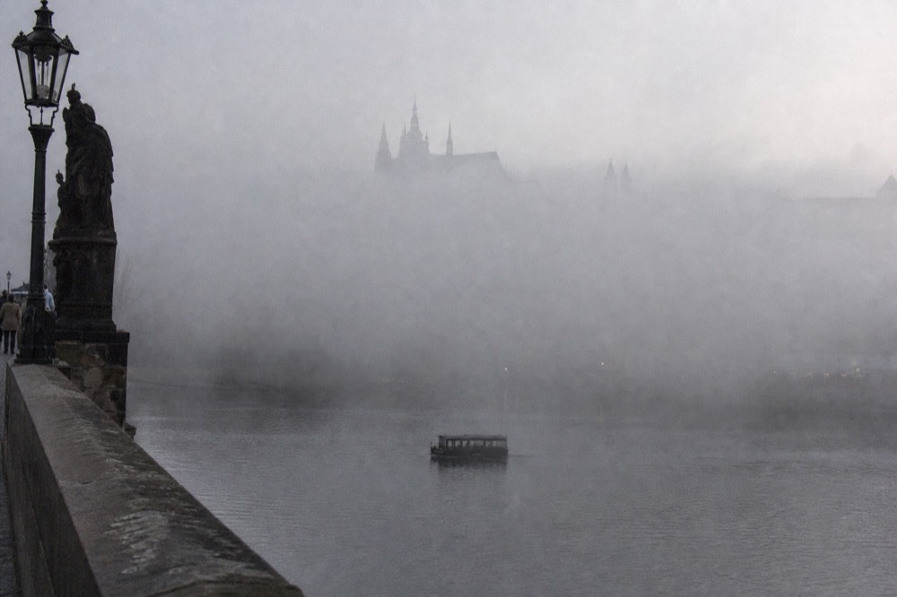 Prague Castle hill wrapped in thick fog, only the cathedral spire breaking through above the mist, the river below barely visible