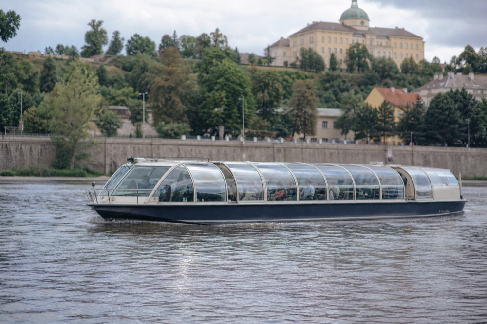 Full exterior view of a covered glass-roof Vltava cruise boat underway, its low unbranded hull and curved glass canopy catching the daylight, the