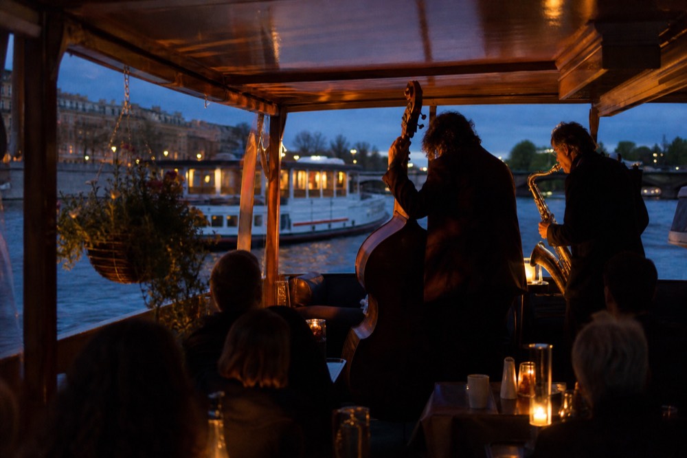 A double bass and saxophone on a small stage on a boat deck, warm stage light against the darkening river