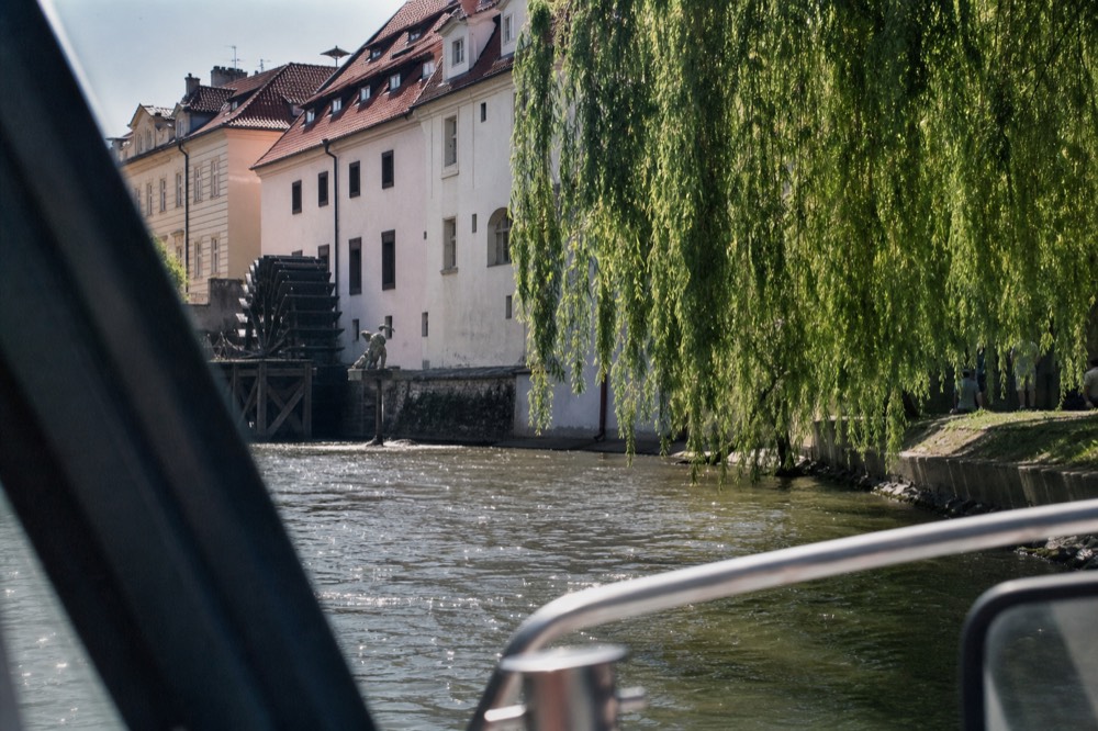 Kampa Island's old waterwheel and riverside park seen from a passing boat, willow branches over the water