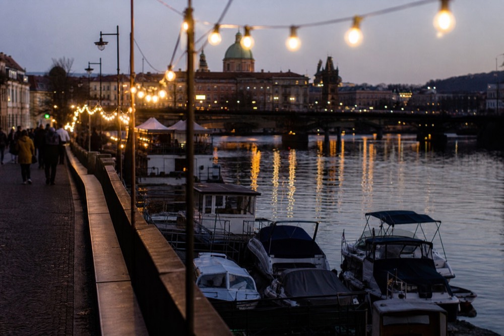 Strings of warm lanterns along a riverside pier at dusk, boats moored below, the water catching the glow