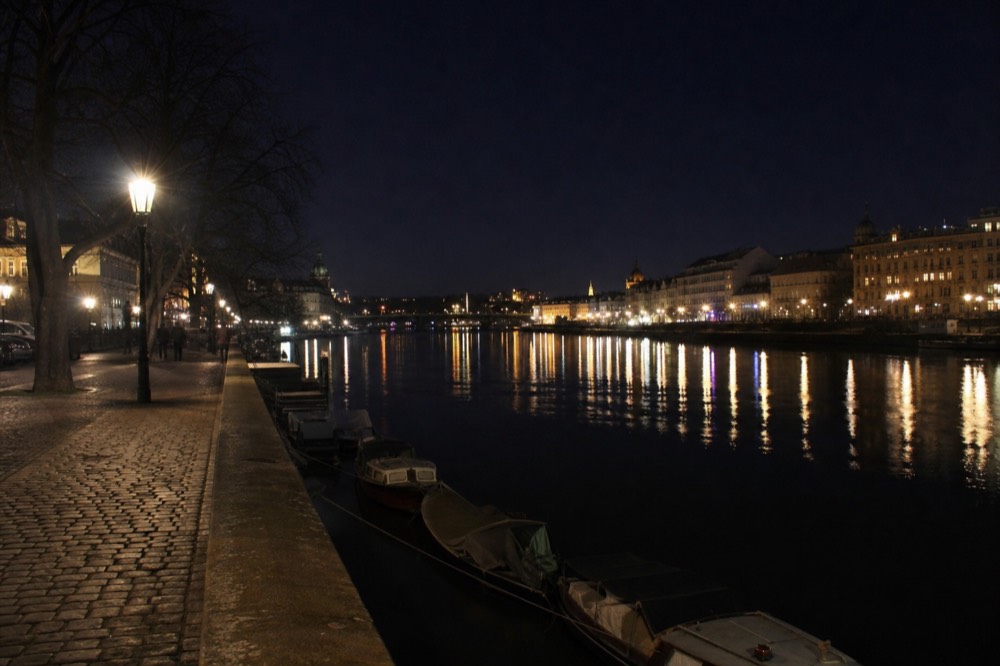 The lit skyline and a near-empty embankment late at night, streaks of lamplight reflected long across the still black water, the sky a deep blue-black