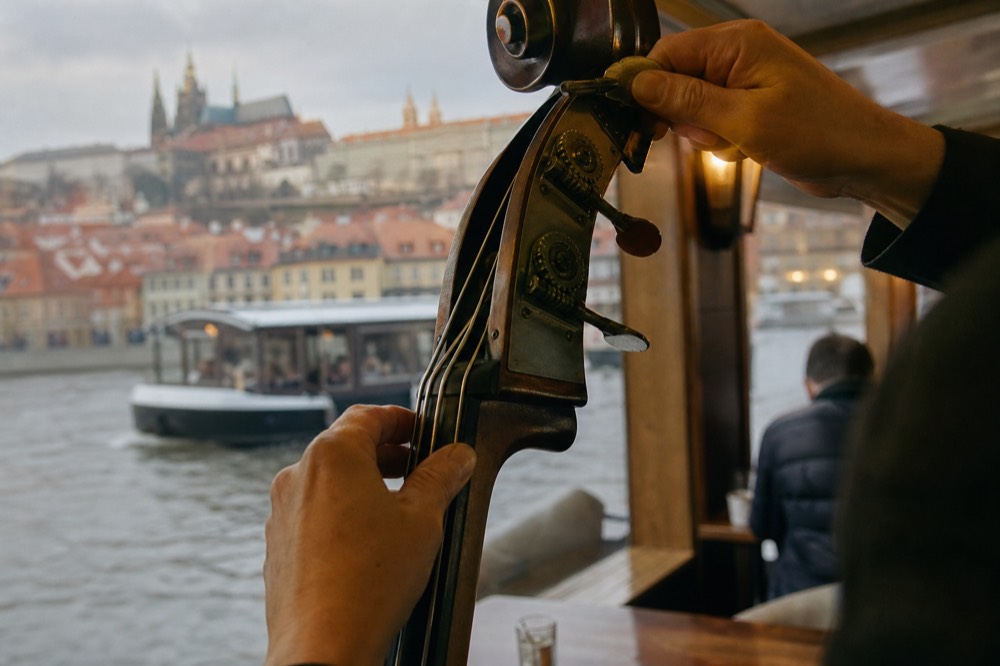 A musician's hands tuning a double bass before a set aboard a Vltava cruise boat, warm cabin light, the instrument's strings and scroll in sharp focus