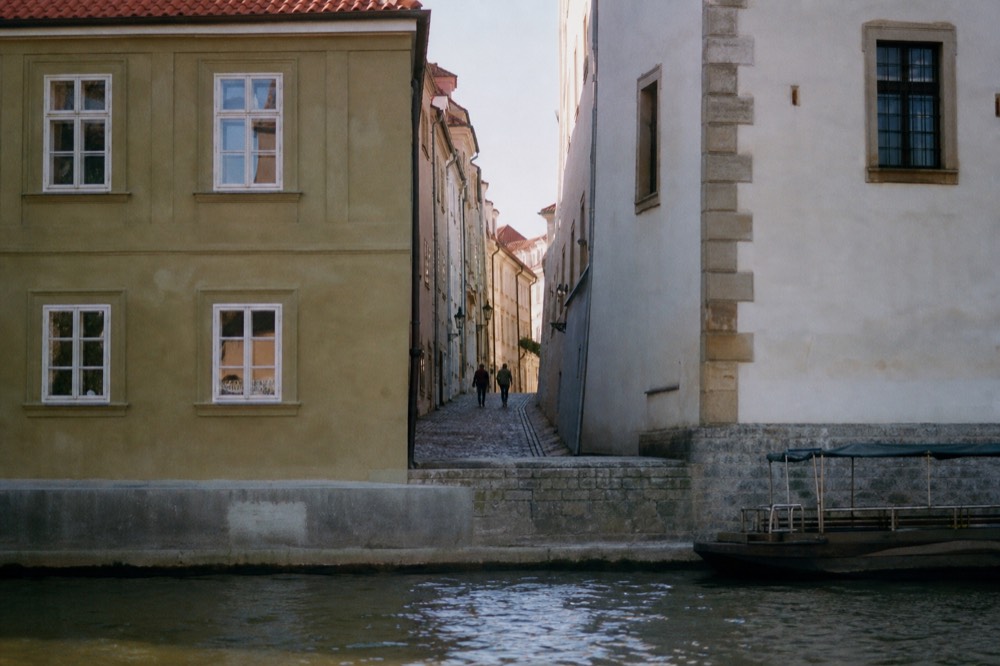 A narrow Mala Strana lane glimpsed from the water between two riverside buildings, sunlit cobbles receding into shadow, the Vltava lapping the foreground