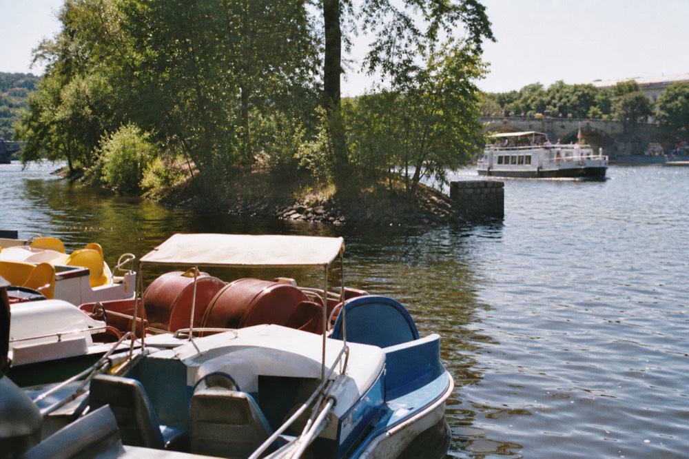 Old-fashioned rental paddleboats bobbing near a small wooded river island, the calm Vltava water dappled with sunlight, a sightseeing boat passing further