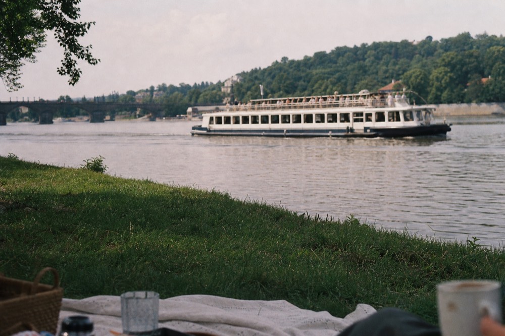A picnic blanket on the grassy Vltava riverbank in summer, a basket and cups just visible at the frame's edge, a sightseeing boat passing across the water