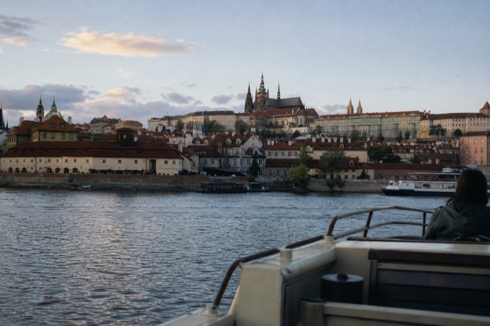 Prague Castle hill in bright daylight seen from a boat deck, red-tiled rooftops stepping down to the river