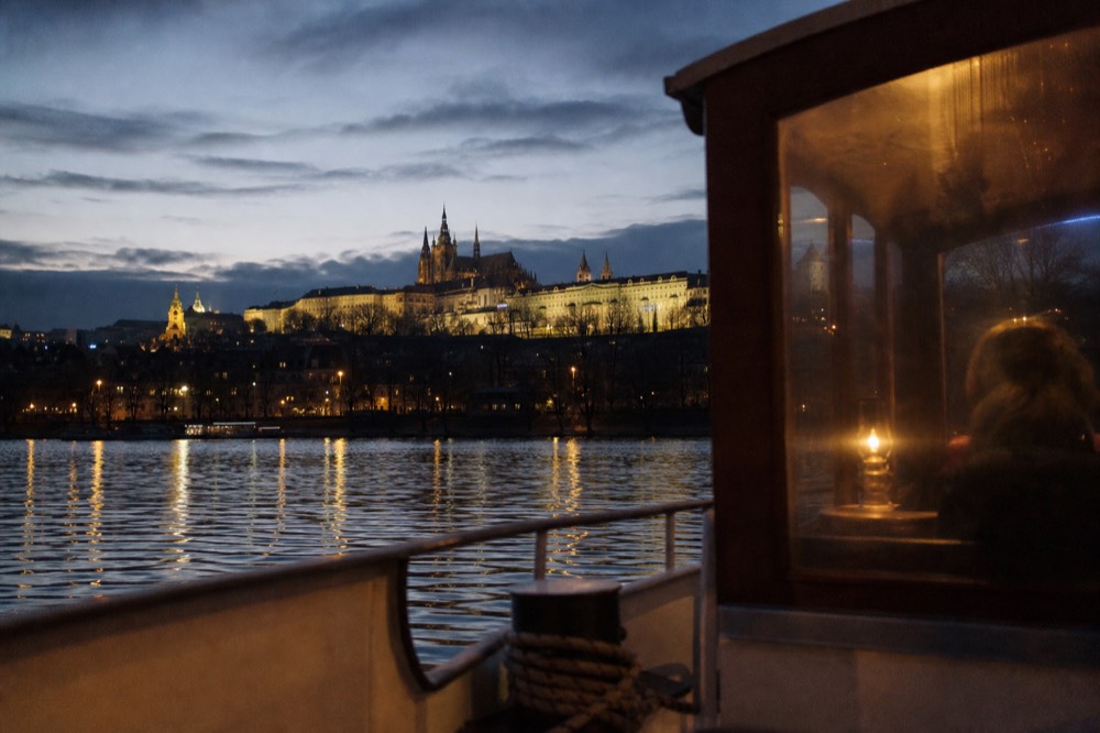 Prague Castle and St Vitus Cathedral in silhouette at dusk, seen across the river from a low boat-deck angle