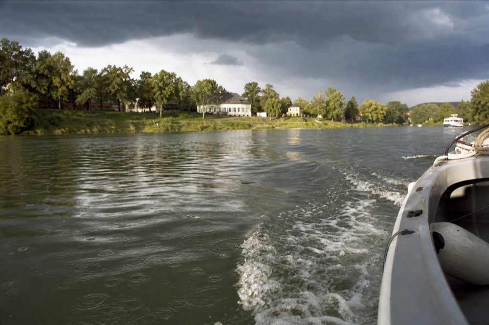 Rain rings spreading across the river surface in soft grey morning light, a boat's wake cutting through