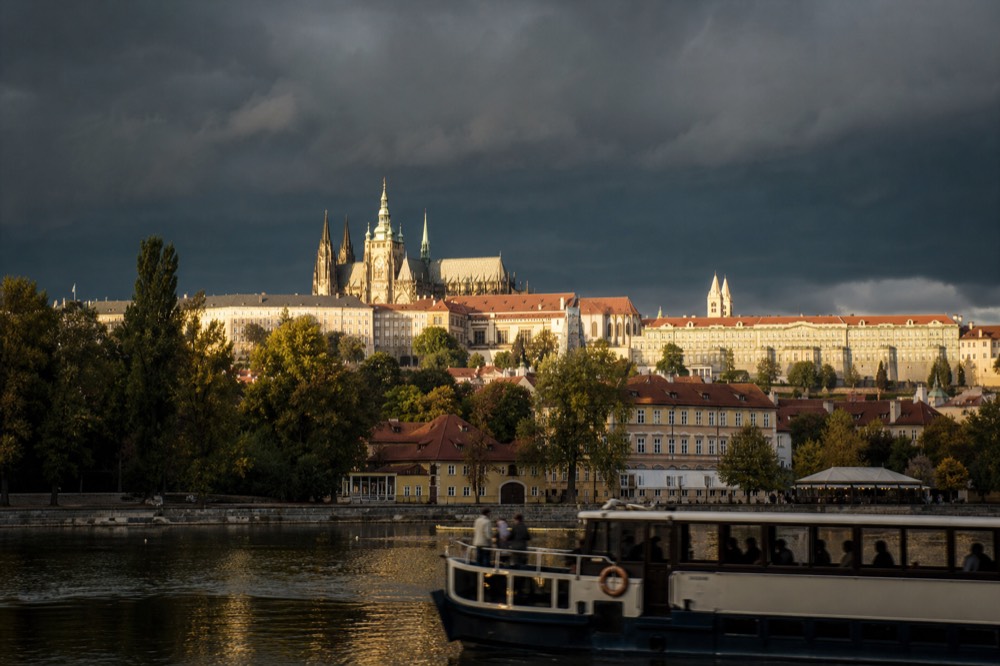 Heavy storm clouds gathering over Prague Castle, a shaft of late-afternoon sun still catching the cathedral spire against the darkening sky
