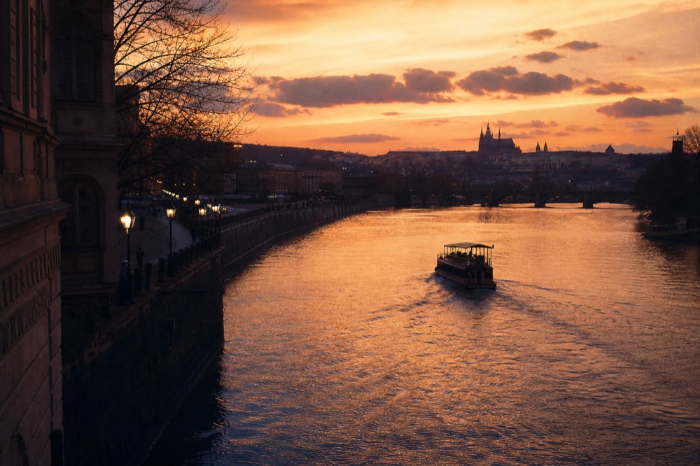 A wide view of the Vltava at sunset, orange sky reflected across the water, one boat's silhouette crossing it