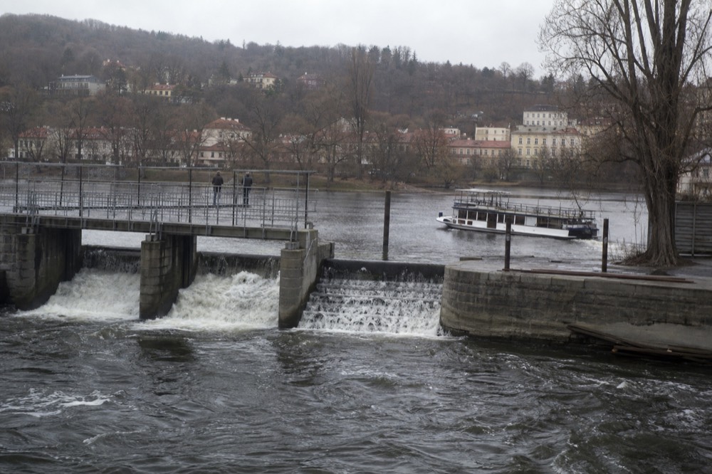 A boat passing through a low weir on the Vltava, white water and spray breaking at the sluice gates