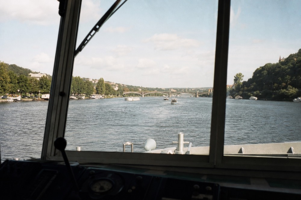 Looking forward through a cruise boat's wheelhouse windshield down the open river, the water and distant bridge framed by the glass and a blurred edge of