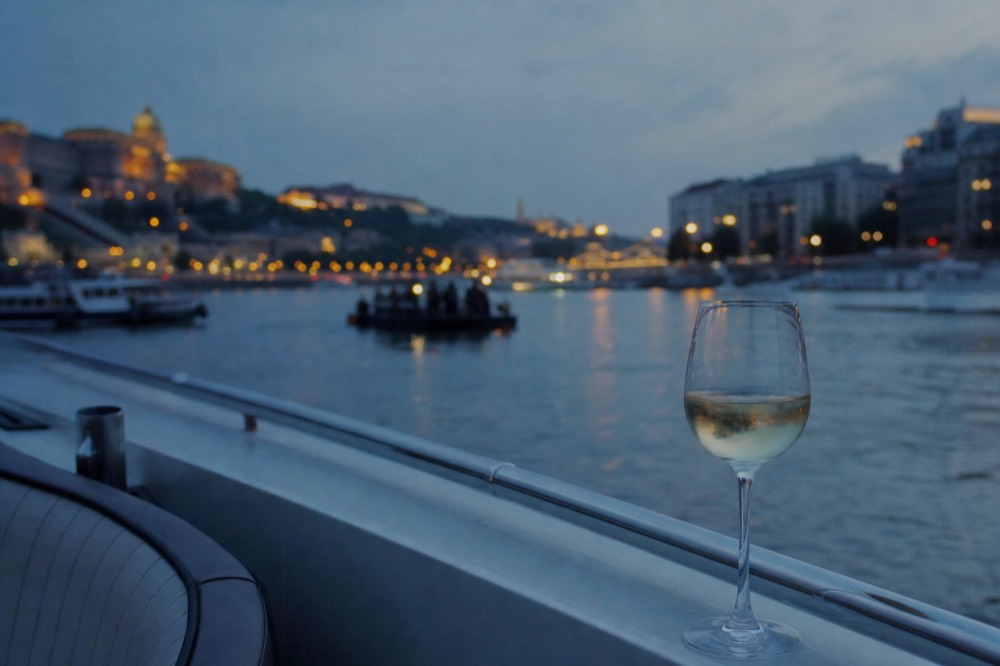 A glass of white wine resting on a boat's deck rail, the lit riverside blurred softly behind it